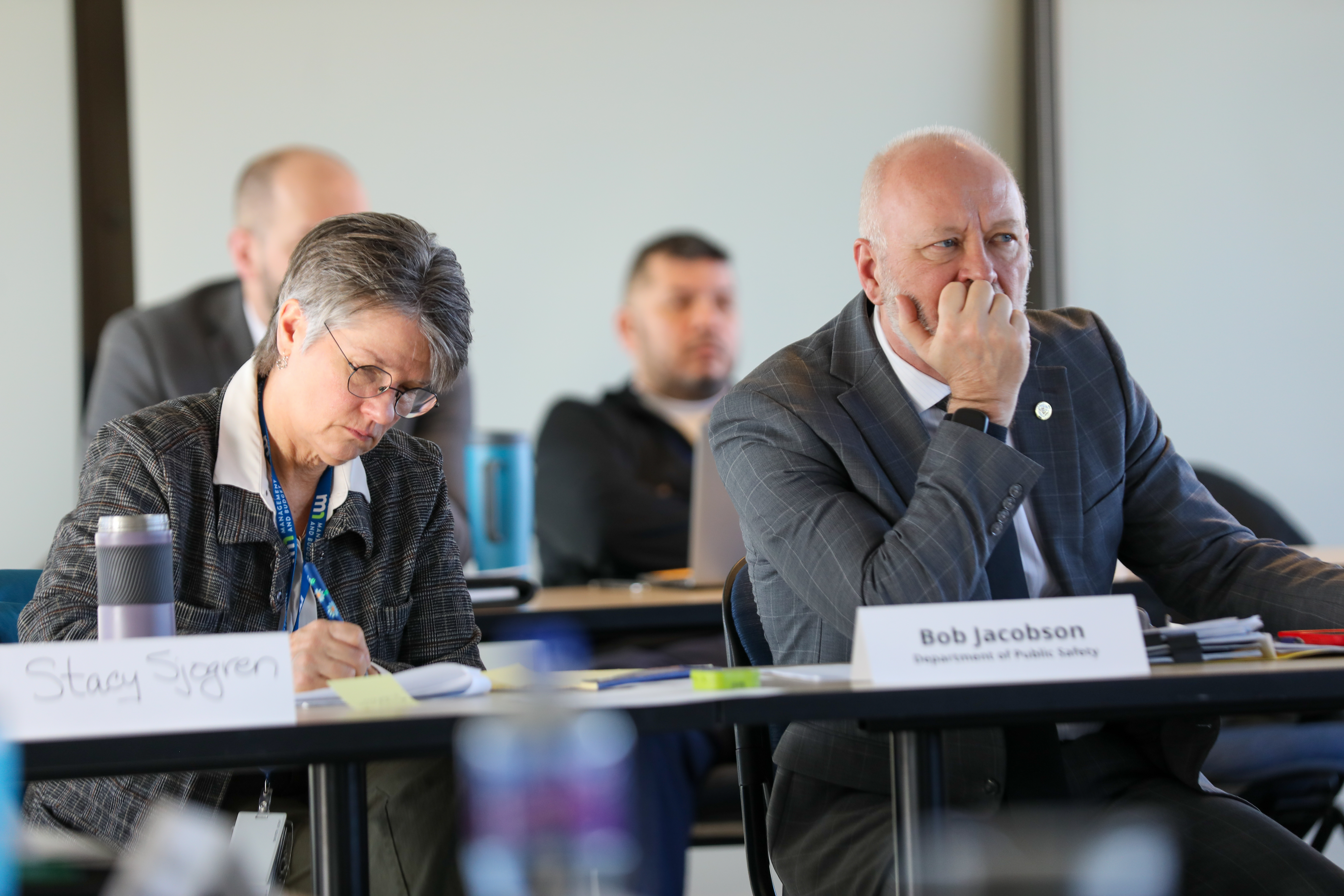 A man in a gray suit and a woman in a gray suit sit at a table taking notes at a meeting.