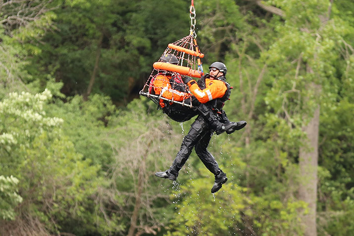 A Minnesota Air Rescue Team member rides inside a rescue basket as another team member hangs to the side while a helicopter lifts them to safety during a water rescue practice.