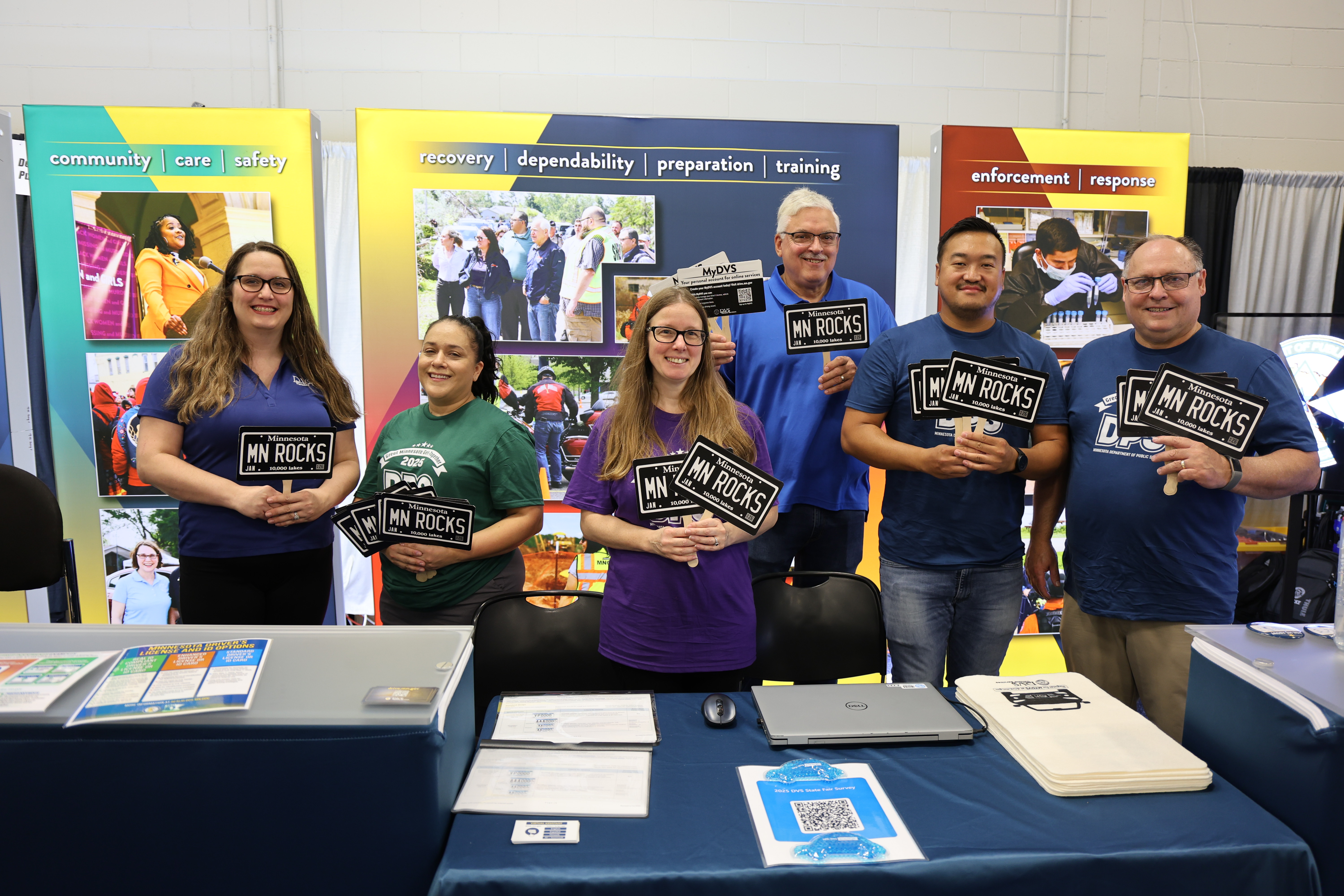 several Department of Public Safety employees behind a table in the Minnesota State Fair DPS booth. They are holding up signs that say MN Rocks.