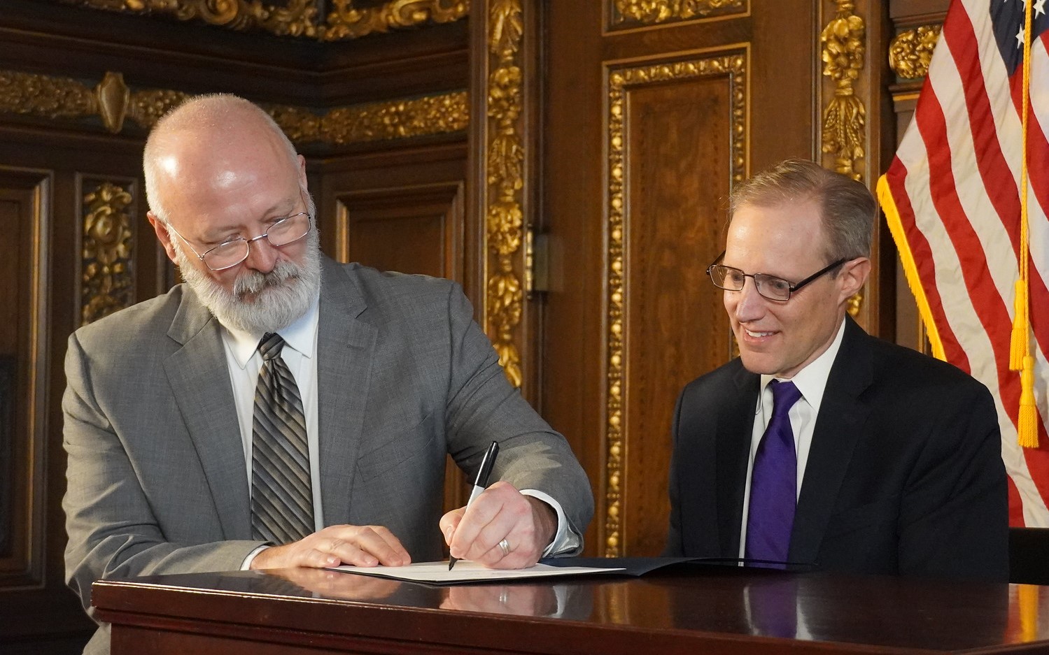 Commissioner Bob Jacobson sits at a desk and signs a piece of paper. 