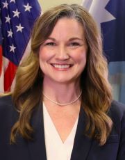 A woman with brown hair smiles while standing in front of the US and Minnesota flags. 