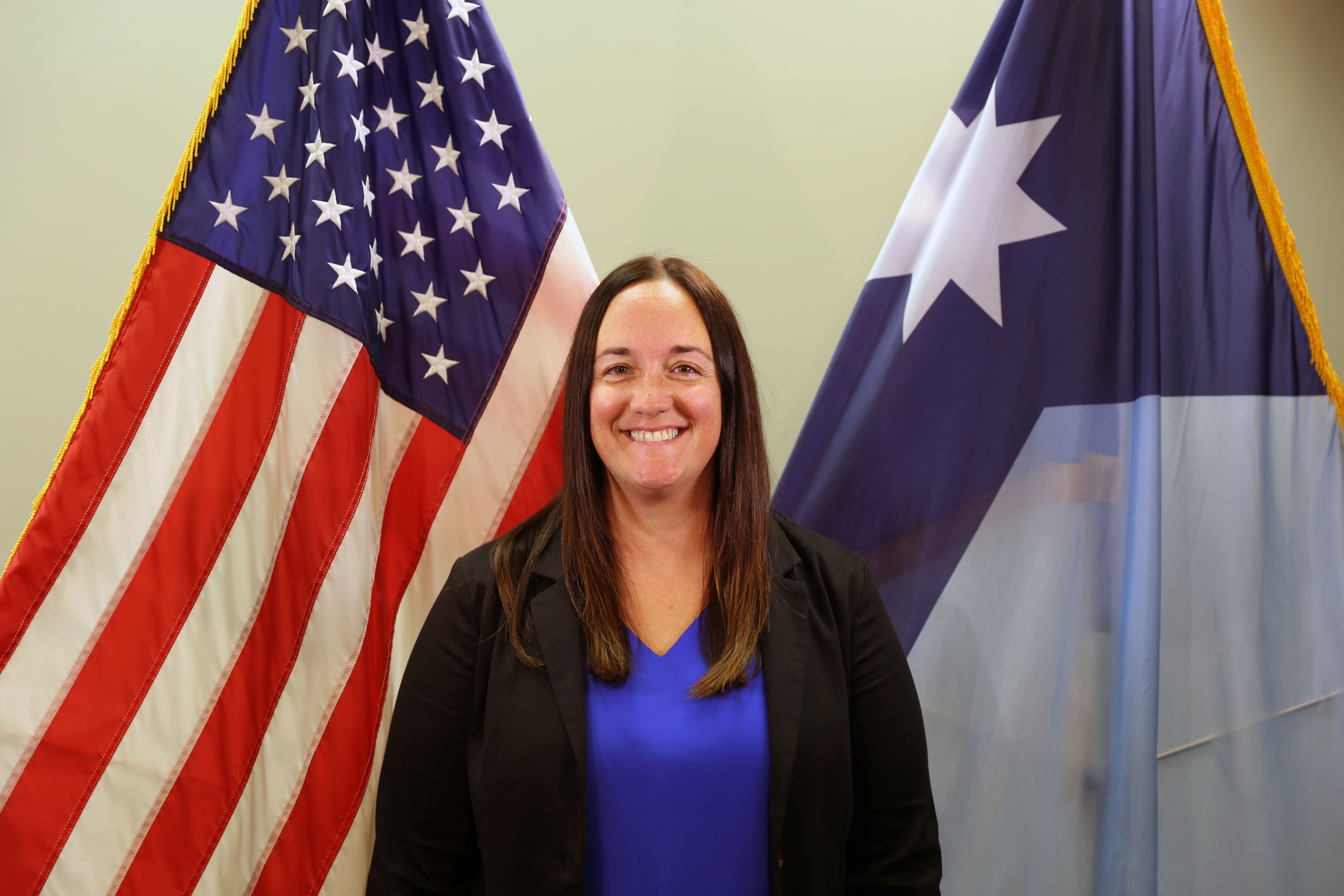 woman in blue shirt and black blazer standing in front of the American Flag and Minnesota State flag