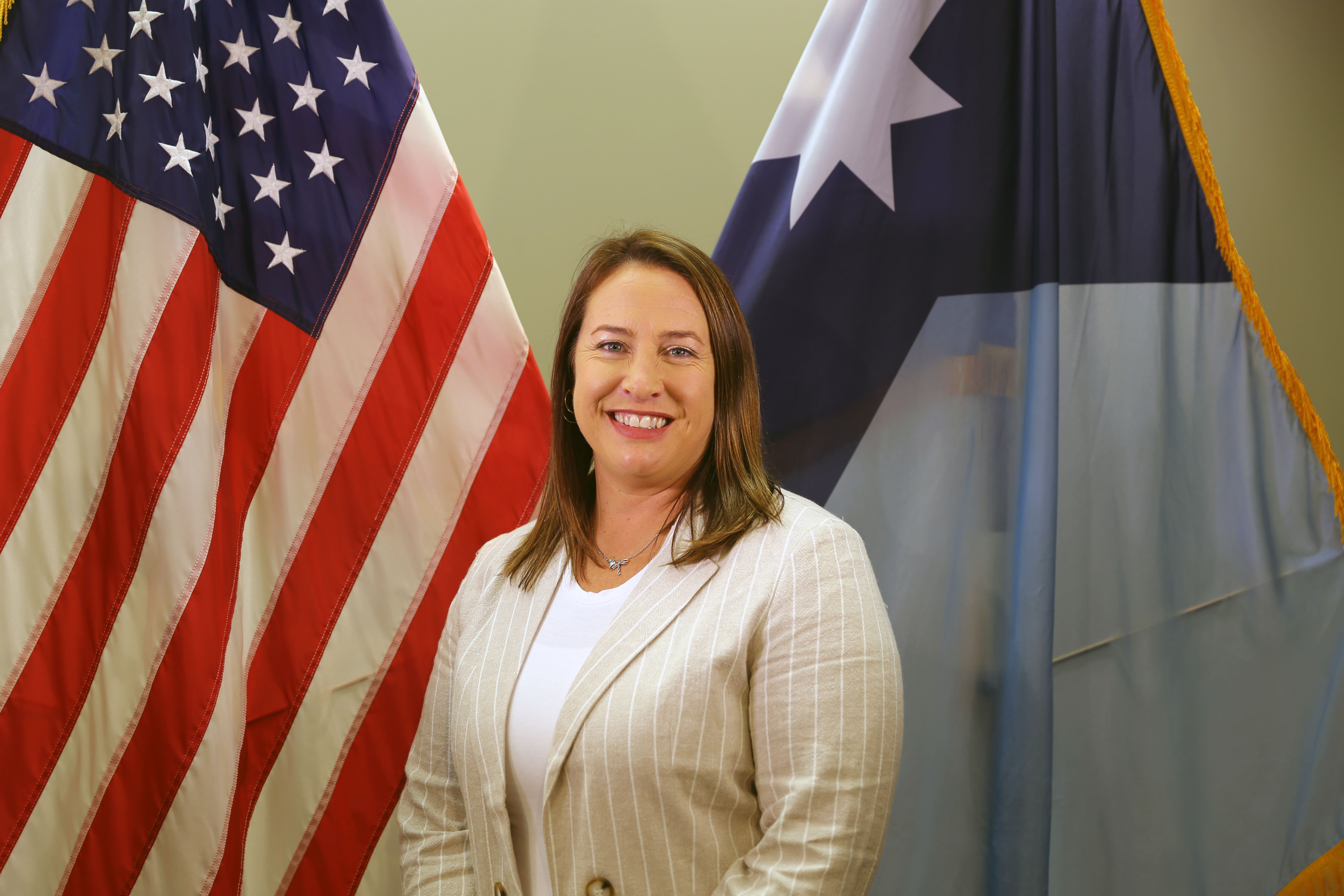 Woman in white shirt and tan blazer standing in front of the American flag and the Minnesota state flag. 