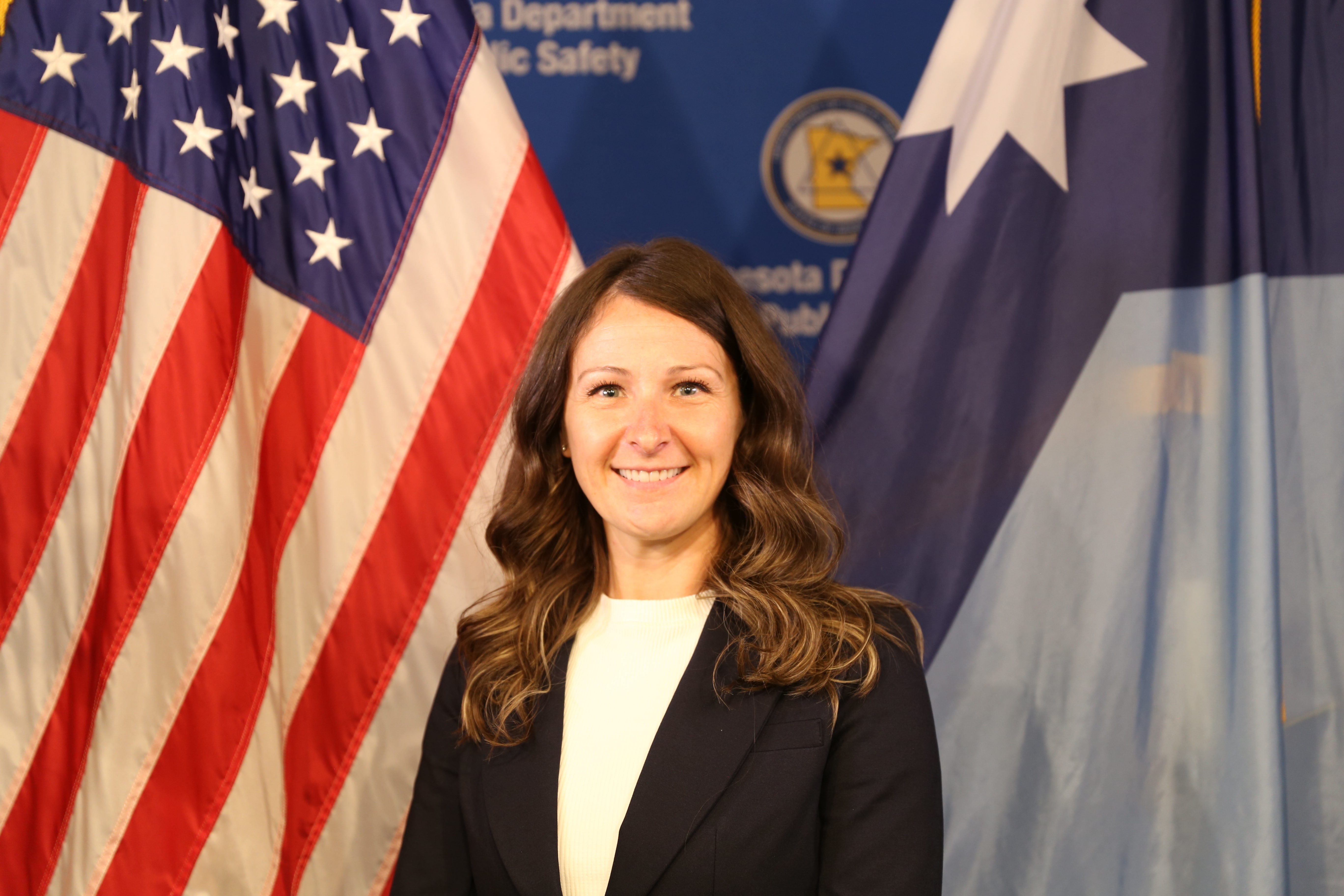 Woman in white shirt and black blazer standing in front of the American and Minnesota flags. 