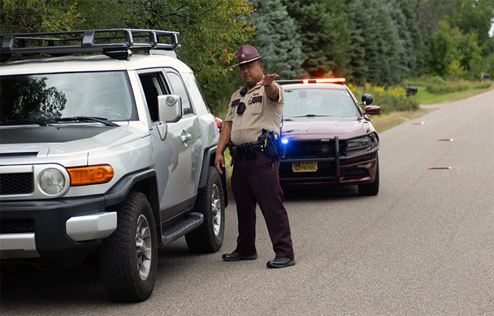A state trooper next to a vehicle on the side of the road, speaking with the driver.