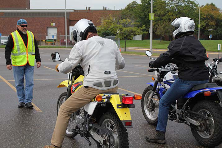Two riders sitting on motorcycles in front of an instructor in a parking lot during a training session.