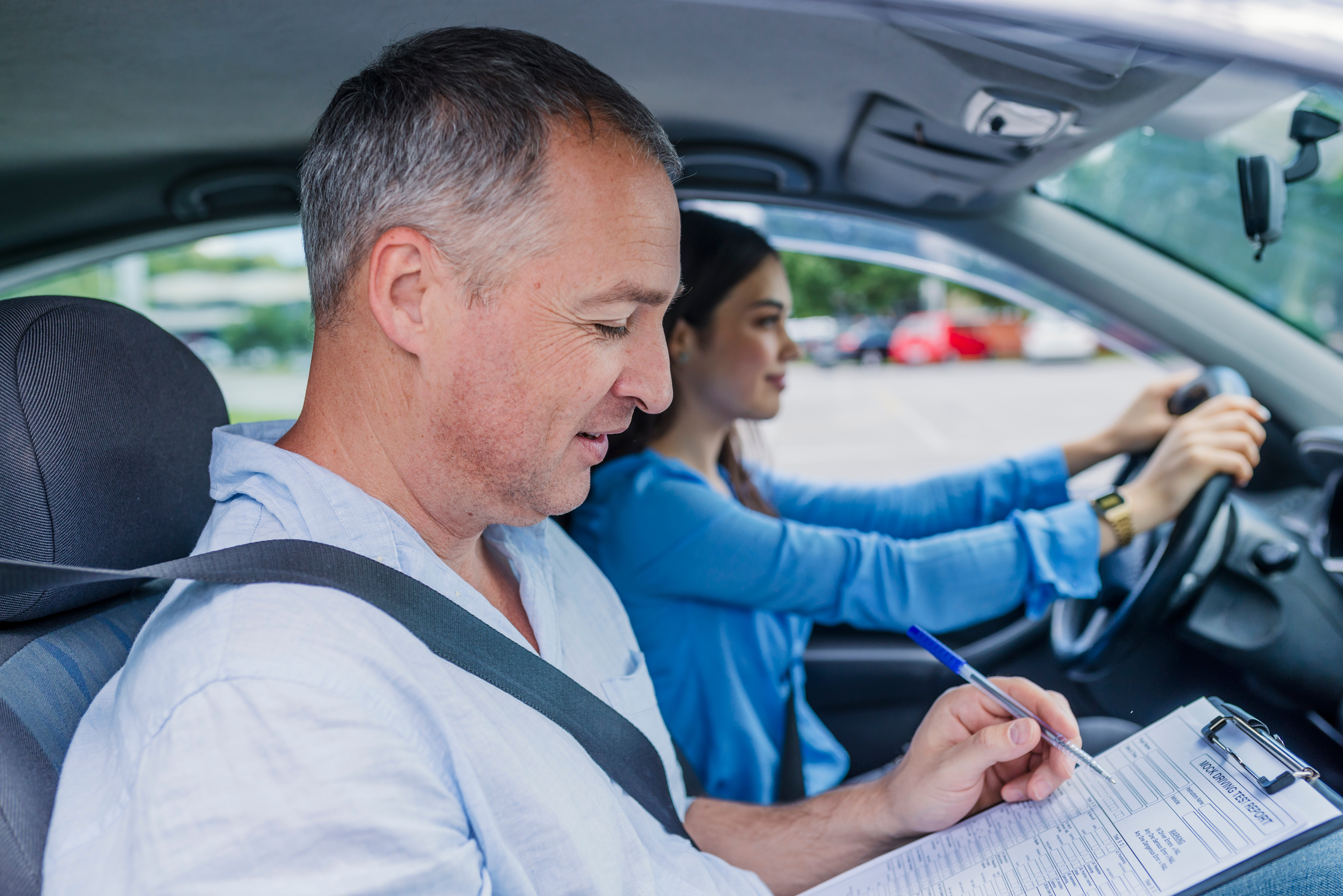 Driver instructor in the passenger seat of a car with a young driver at the wheel. The instructor is writing on a clipboard.